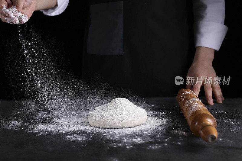 厨师用手将面粉粉倒入生面团上，用筛子在黑色的背景上，烹饪过程