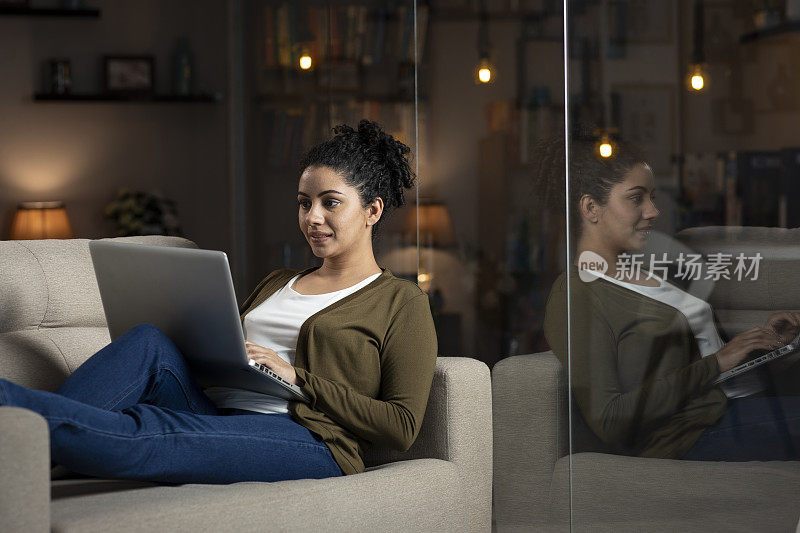 一个年轻女子坐在靠近玻璃窗的沙发上用笔记本电脑上网的照片