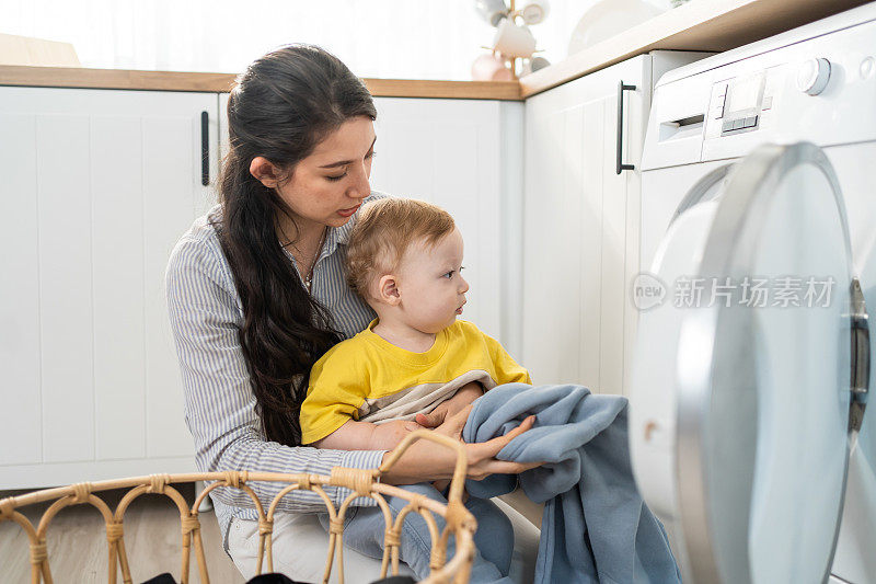 白种人忙碌的母亲做家务与婴儿男孩蹒跚学步在厨房。漂亮的年轻妈妈把衣服放进洗衣机，而她的小儿子在家里玩。Family-housekeeping。