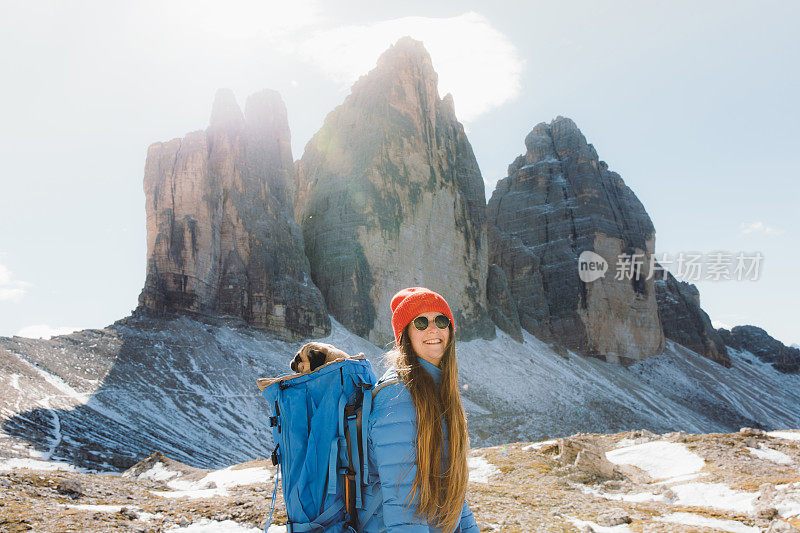 年轻女子抱着狗在背包里凝视风景优美的山脉徒步旅行在白云石阿尔卑斯山