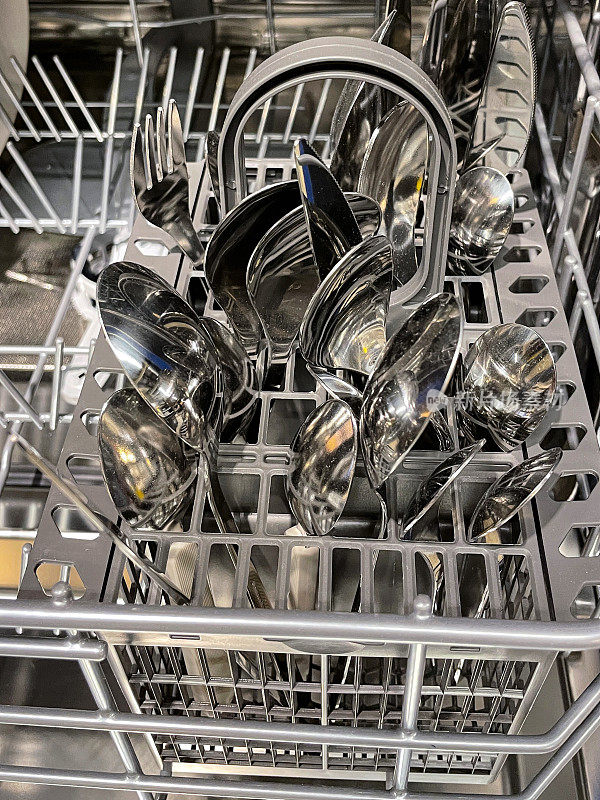 塑料洗碗机内部的特写图像，餐具篮里装着干净的不锈钢刀、叉和勺子，机器半满，干净的餐具在清洗和干燥后可以收起来