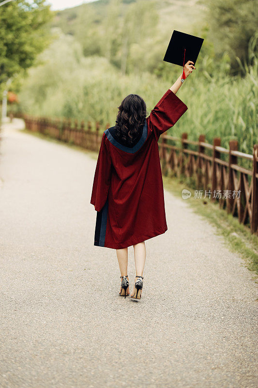 绿色公园里身穿毕业礼服的年轻女子手持帽子的背影