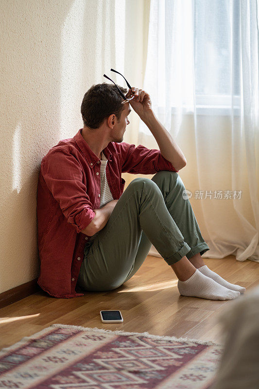 一名沮丧的欧洲男子拿着手机坐在自家的地板上，悲伤地望着窗外