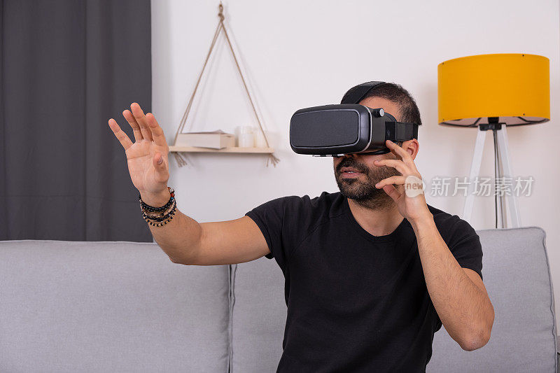 年轻男子戴着VR眼镜坐在家里的沙发上兴奋地打手势，未来科技，教育视频游戏