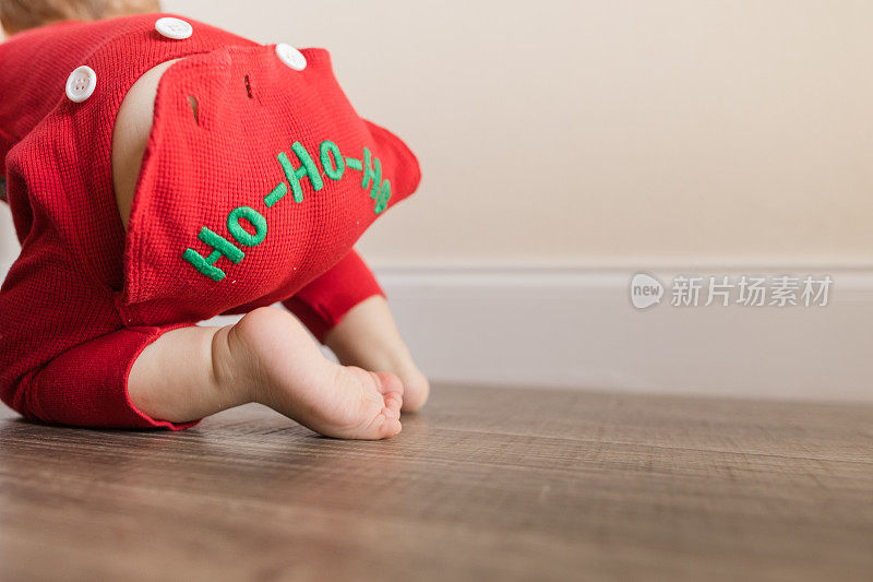 一个11个月大的圣诞男婴长着12只脚趾，穿着红色复古风格的圣诞睡衣，睡衣底部写着“HO-HO-HO”，在木地板上玩耍，为他的生日和2022年的假期做准备