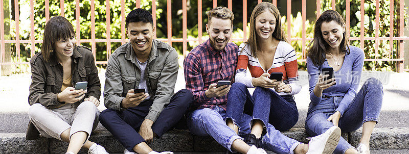 △在城市的人行道上，多文化的朋友们用智能手机开怀大笑的横横幅或标题:沉迷于手机的年轻人