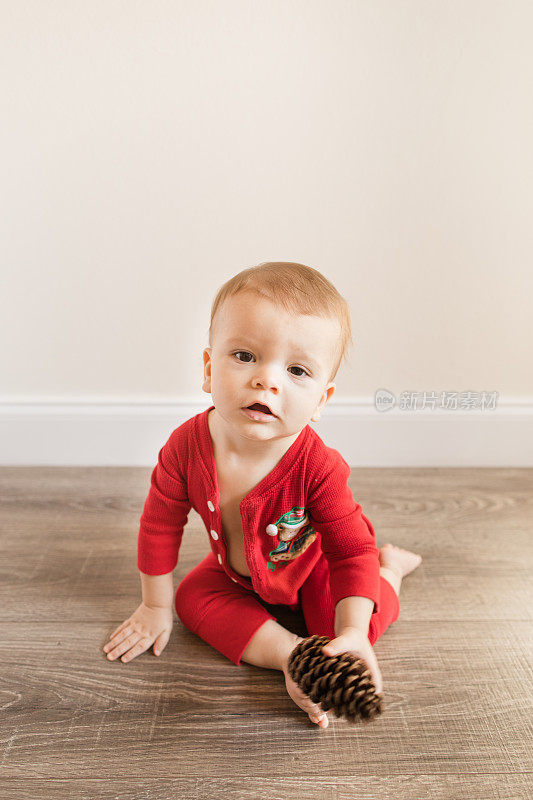 一个11个月大的圣诞小男孩穿着红色复古风格的圣诞睡衣，前扣敞开着，在木地板上玩松果，为他的生日和2022年的假期做准备