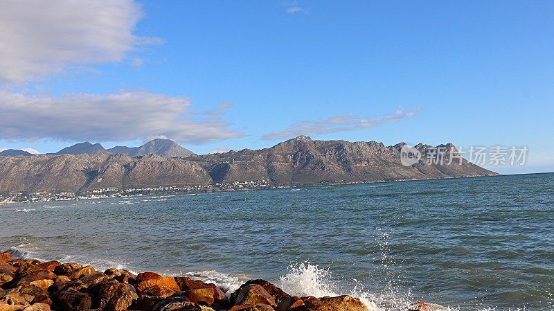 南非开普敦，海浪撞击着海滩上的岩石，背景是戈登湾的山脉