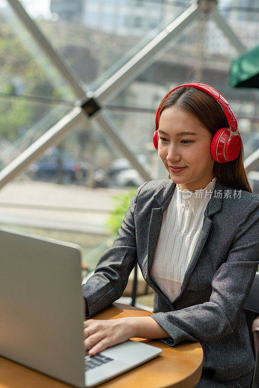 混合式工作风格。年轻的亚洲女性成年人与笔记本电脑在咖啡馆工作。戴着红色耳机的女商人