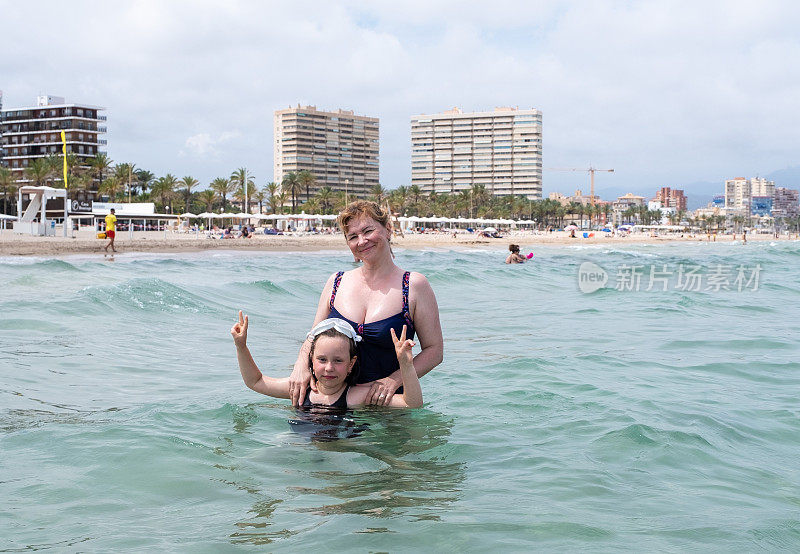 一个女人和一个7岁的女孩，白种人在海里游泳。在沙滩上休息。阿利坎特海岸部分和建筑。