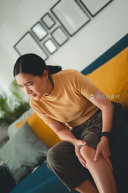 一位亚洲妇女坐在家里客厅的沙发上忍受着膝盖疼痛。