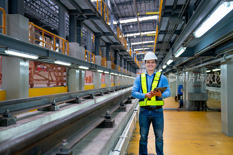 专业工程师工人的肖像拿着平板电脑，微笑着看着相机，也站在工厂工作场所的铁轨旁。