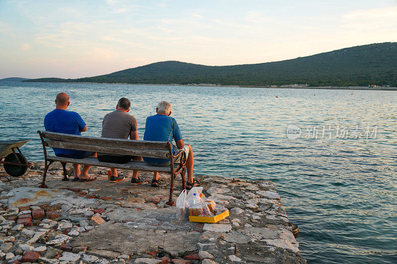 克罗地亚亚得里亚海的小镇上，三个人坐在长凳上，与蓝天、碧海、多云的天空、航行的寂静和啤酒的盒子一起演奏交响乐。