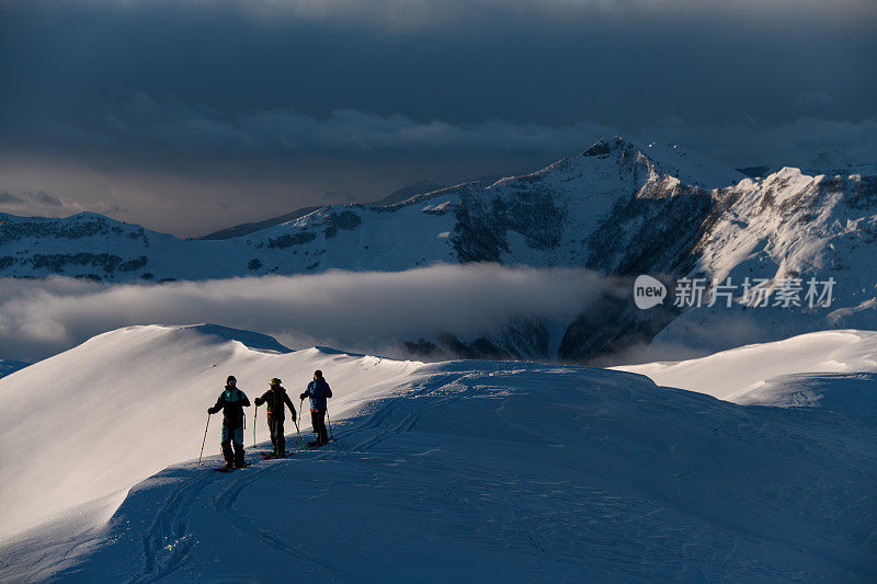 三名男性滑雪者站在一座白雪皑皑的山峰上，山峰只有一部分被阳光照亮