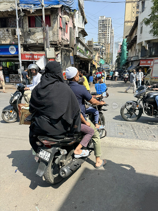 两个无法辨认的人坐在零售区的助力车上的特写图像，女乘客穿着朴素的衣服，男司机戴着头巾，没有防撞头盔，零售商店，后视图，重点在前景