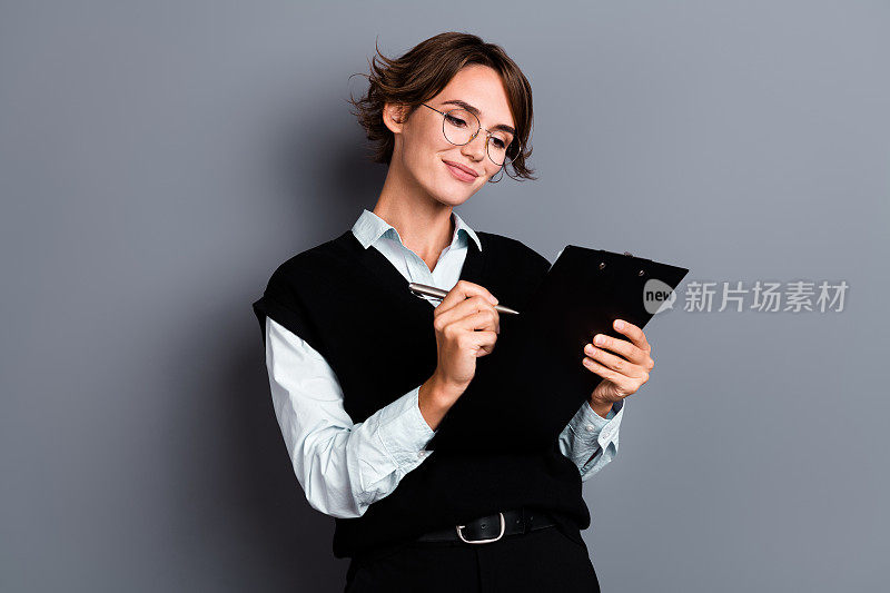 照片中，成功的女企业家穿着时髦的衣服，拿着纸架，在灰色背景上写笔记，每周计划