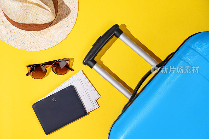 黄色背景上平躺着一个蓝色手提箱，里面装着旅行用品、帽子和太阳镜。