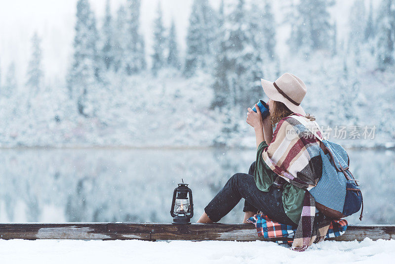 拿着热饮料和油灯的女人坐在白雪覆盖的湖边