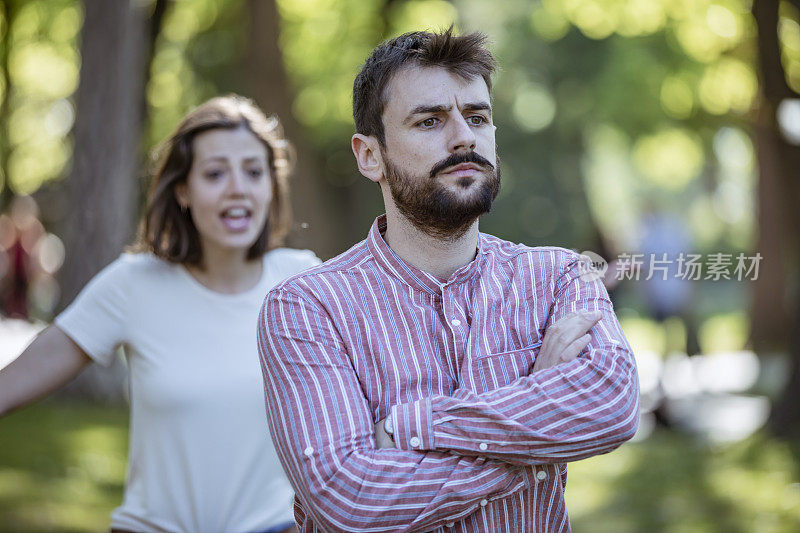 一对年轻夫妇在公园里吵架。年轻夫妇有一个夏天的一天争吵。站在公园外面，困惑的年轻女子打着手势，看着她的男朋友