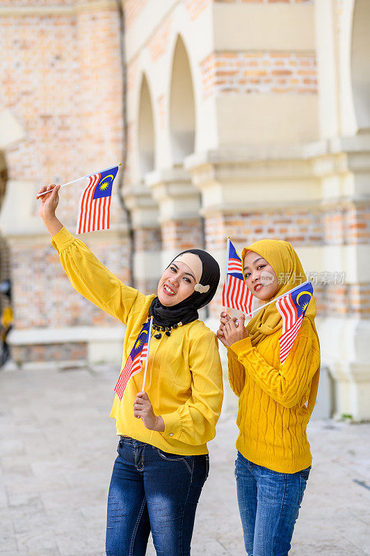 两个年轻漂亮的穆斯林妇女戴着头巾看着镜头和马来西亚国旗