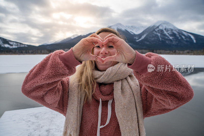 一名年轻女子在雪山松林上制作心形手指架，享受寒假的乐趣