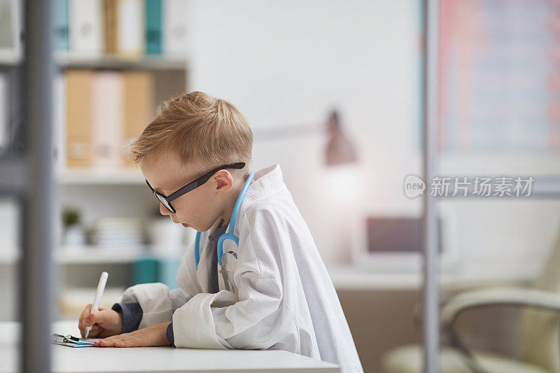 严肃专注的小男孩穿着医生的衣服站在桌子旁，在医疗档案中做笔记