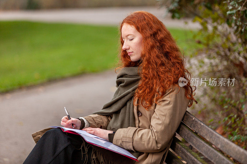 一个年轻女子坐在长椅上写作业，这是城市公园里的宁静场景