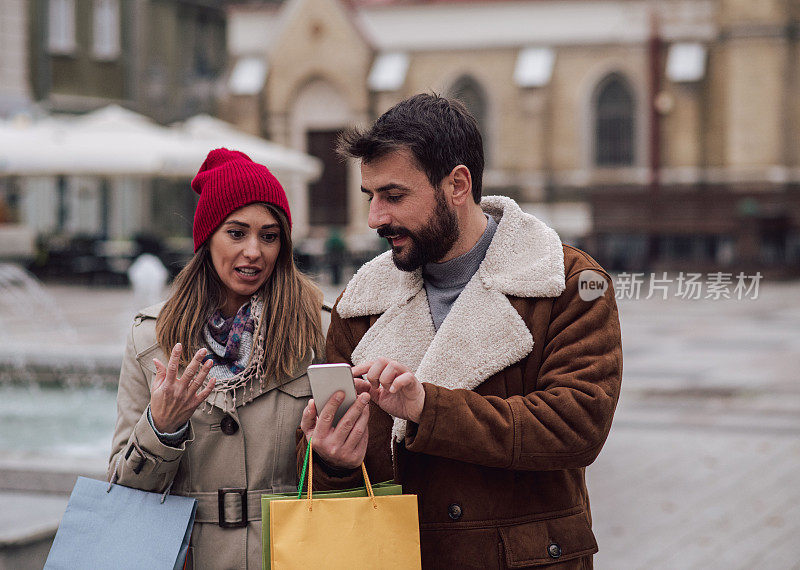 一个年轻的女人正在看她男朋友拿着的手机。他给她看令人不安的消息。市中心的购物日。寒冷的秋日。