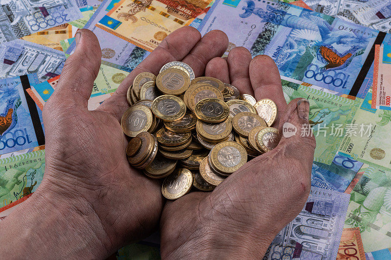 一名工人的双手拿着一堆100坚戈和200坚戈面额的硬币，背景是哈萨克斯坦钞票