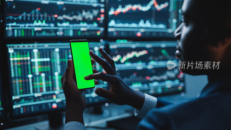 金融分析师使用智能手机与绿色屏幕色度键模拟模板和工作在多显示器工作站与股票图表。商人晚上在投资银行工作。