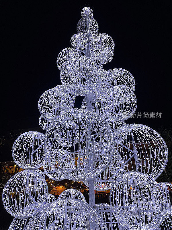 夜晚的特写图像，白色LED仙女灯覆盖的球体堆叠在一起，形成巨大的户外圣诞树，黑暗的天空