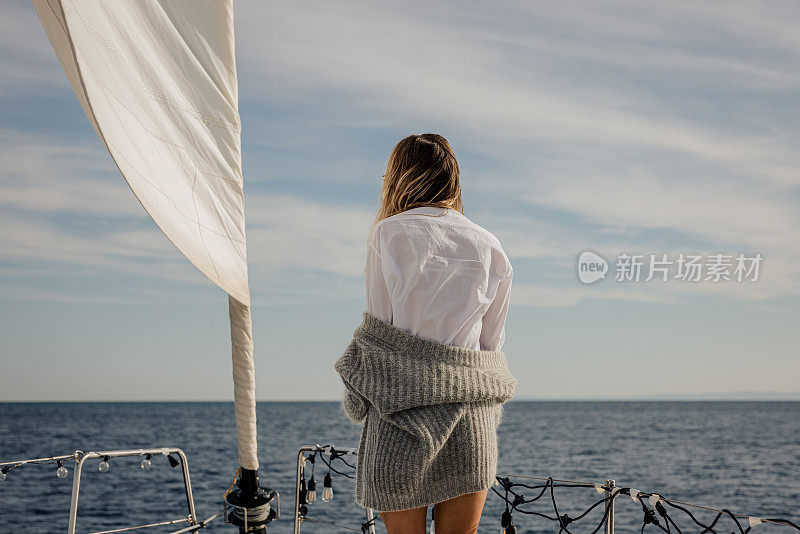 长发女子，穿着灰色套衫站在帆船的船头甲板上，后视图，大海在背景中