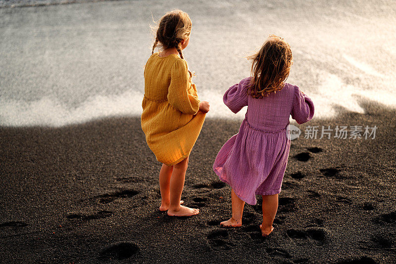 快乐可爱的小女孩们在沙滩上享受阳光明媚的日子，站在海岸上看着海浪。家庭暑假。姐姐的爱。