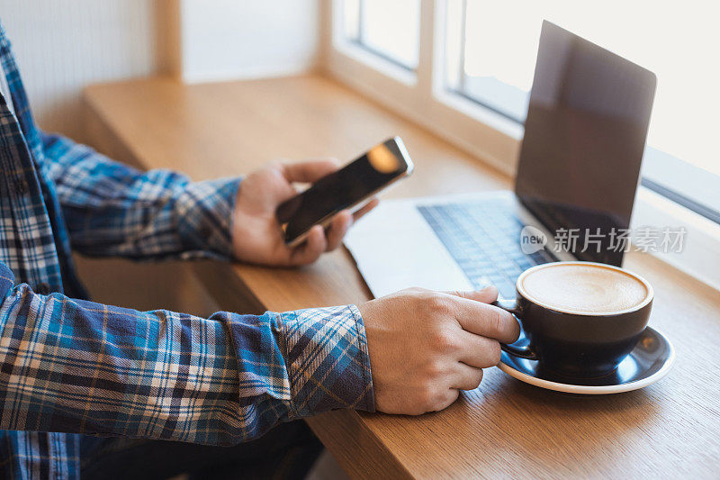 一个男人坐在咖啡馆里，端着一杯咖啡，一边用笔记本电脑和智能手机工作
