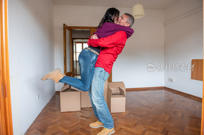 穿着牛仔裤的丈夫抱着被纸板箱包围的妻子，兴奋地搬到新公寓，快乐的白人夫妇愉快地拥抱，一起搬到自己的新家。