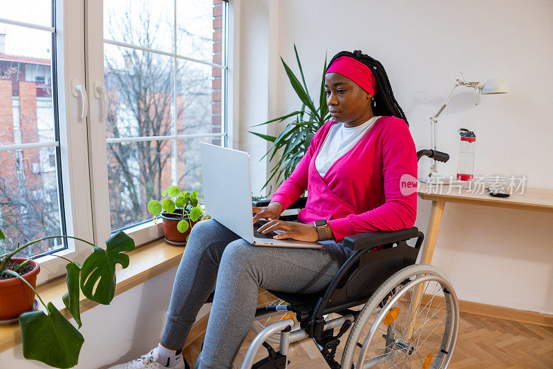 年轻的黑人妇女坐在轮椅上用笔记本电脑工作，靠窗