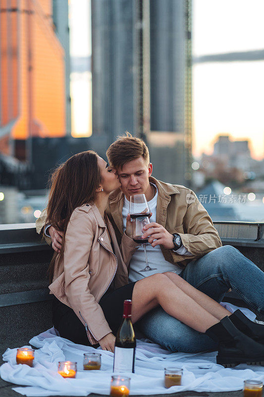 以城市景观和摩天大楼为背景的屋顶惊喜约会。幸福的年轻情侣喝着酒，吃着浪漫的烛光晚餐，庆祝周年纪念日或情人节