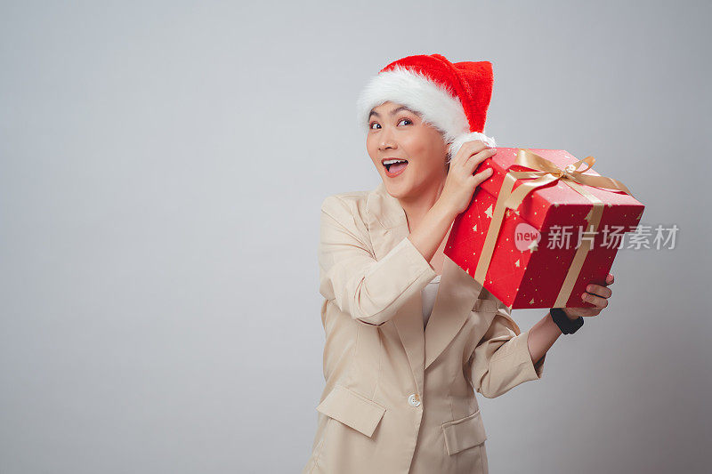 亚洲女人戴着圣诞老人的帽子开心地微笑着摇着礼盒，孤立在白色的背景上。