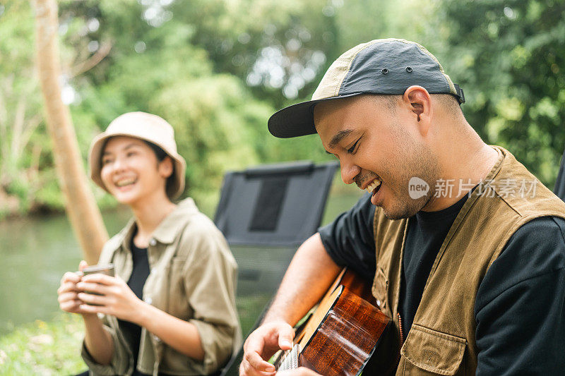 年轻的亚洲夫妇，男人和女人野餐坐在露营地户外花园。两个人在森林里露营，弹吉他，喝咖啡。旅游，放松，露营，度假，假日，周末的主题