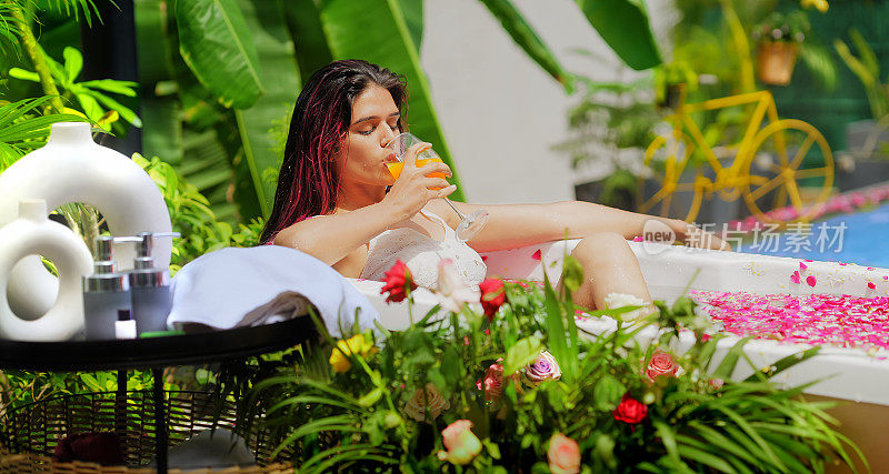 印度微笑的z世代女性坐在浴缸里放松，喝着健康的新鲜果汁。美丽的年轻成年女孩早上沐浴玫瑰花瓣水享受暑假在酒店度假别墅之旅