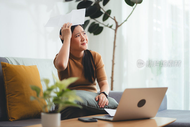 亚洲女性坐在沙发上，担心银行票据、抵押贷款债务和生活成本的账单支付问题。