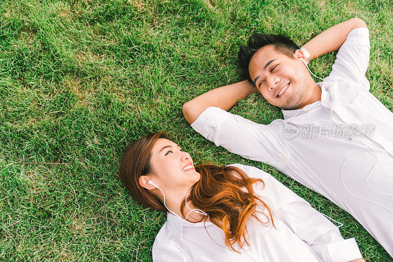年轻的亚洲可爱的情侣或大学生一起躺在草地上，听着音乐，顶视图与抄写空间。爱情，关系，婚礼，或放松休闲的生活方式概念