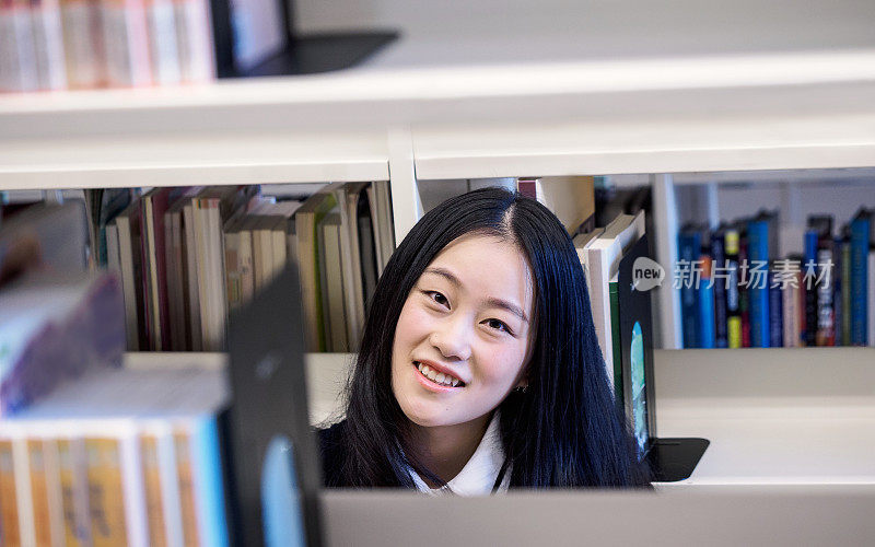 一个美丽的中国学生在大学图书馆的书架后面微笑的大头照，图书馆和教育理念。