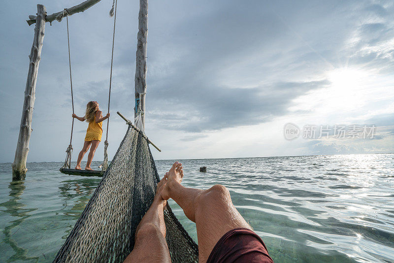 年轻夫妇在海边的海滩上荡秋千，美丽的田园诗般的风景。人们旅行浪漫度假的概念。男人在吊床上和女朋友在海上荡秋千的个人观点。