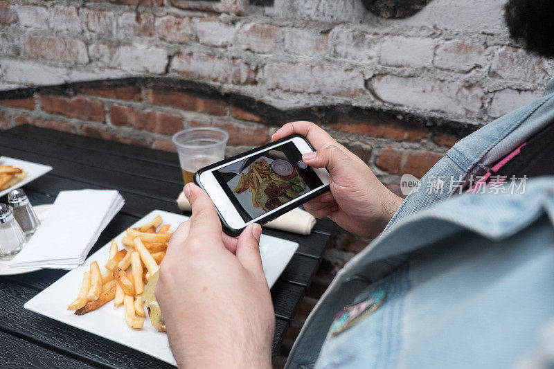 一名男子拿着手机给他的食物拍照。用手机拍摄食物。用智能手机给户外酒吧里的汉堡、啤酒和炸薯条拍照。