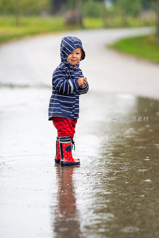 小女孩穿着雨衣站在乡村道路上的水坑里