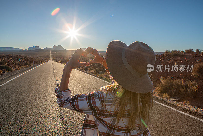 爱美国的女人，把手指做成心形;女人的后视图站在漫长的高速公路通往纪念碑谷在美国。人们在美国旅行的概念