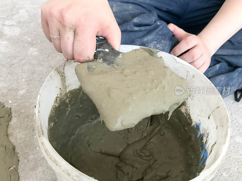 建筑工手用金属刮刀在一个大的白色塑料建筑桶中揉捏灰泥、瓷砖胶水、用于修复公寓、房屋的水泥、找平墙壁和浇筑平整面