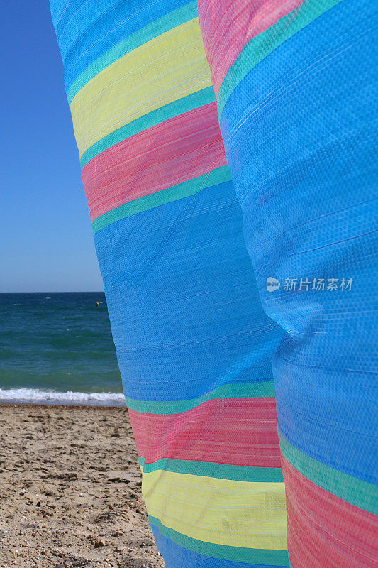色彩鲜艳的条纹防风林在沙滩上随风翻滚，背景中海浪拍打着水的边缘，聚焦于前景
