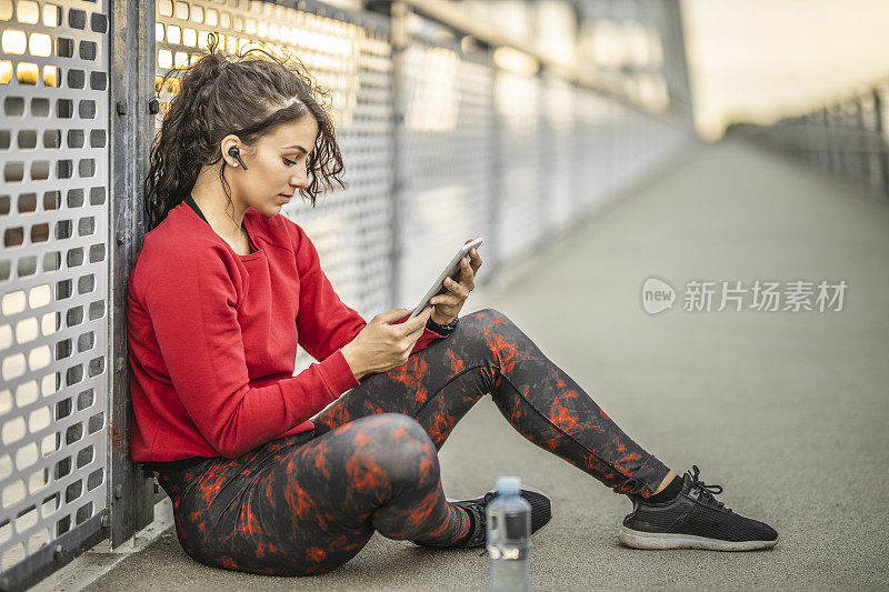 年轻的运动女性在户外艰苦训练后休息，听音乐，使用数码平板电脑和喝水